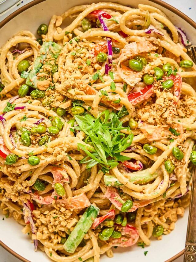 A top-down view of a bowl of sesame noodle salad, filled with creamy noodles, roasted edamame, red bell pepper, carrots, purple cabbage, and garnished with crushed peanuts, sesame seeds, and fresh green onions. Two silver forks rest on the side of the bowl.