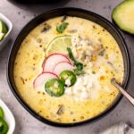 A black bowl filled with creamy Chicken Poblano Soup, garnished with sliced radishes, jalapeños, lime, cilantro, and crumbled cheese. A spoon rests in the bowl, and slices of avocado and lime are visible in the background.