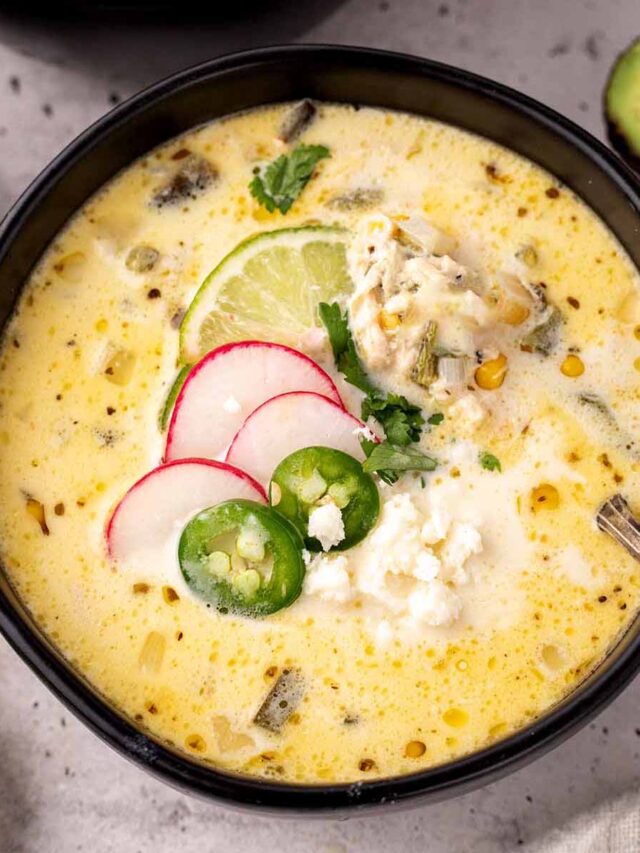A black bowl filled with creamy Chicken Poblano Soup, garnished with sliced radishes, jalape&ntilde;os, lime, cilantro, and crumbled cheese. A spoon rests in the bowl, and slices of avocado and lime are visible in the background.