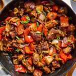 Close-up of chili crisp chicken stir-fry featuring golden-brown chicken, red bell peppers, onions, and scallions, coated in a glossy chili crisp sauce and garnished with fresh herbs, served in a skillet.