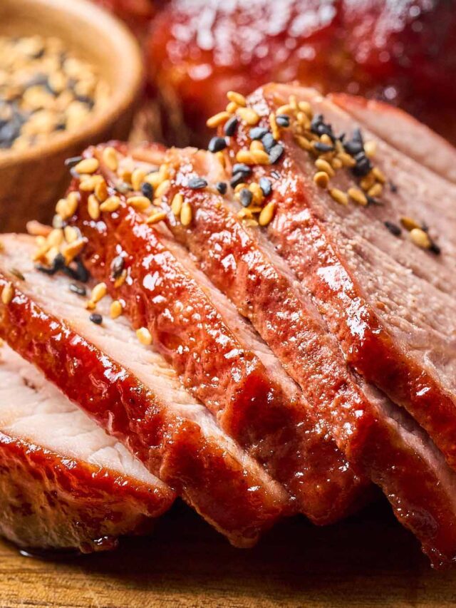 Close-up of five-spice pork slices, garnished with sesame seeds and served on a wooden board, with a small bowl of sesame seeds in the background.