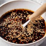 Garlic sesame sauce in a white bowl, topped with toasted sesame seeds and red pepper flakes, with a wooden-handled spoon resting inside.