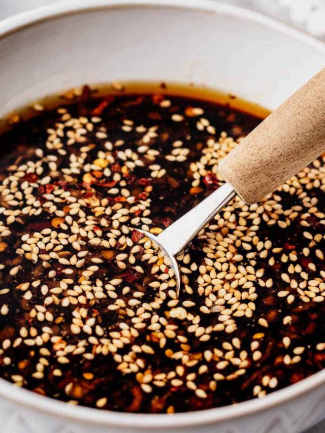 Garlic sesame sauce in a white bowl, topped with toasted sesame seeds and red pepper flakes, with a wooden-handled spoon resting inside.