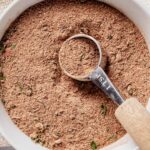Close-up of homemade jerk seasoning in a white bowl with a 1 teaspoon measuring spoon resting on top, showcasing the textured spice blend with flecks of dried herbs.