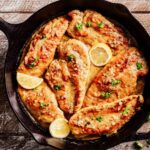 Overhead view of Limoncello Chicken in a cast iron skillet, featuring golden seared chicken breasts coated in a creamy citrus sauce, garnished with fresh parsley and lemon slices.