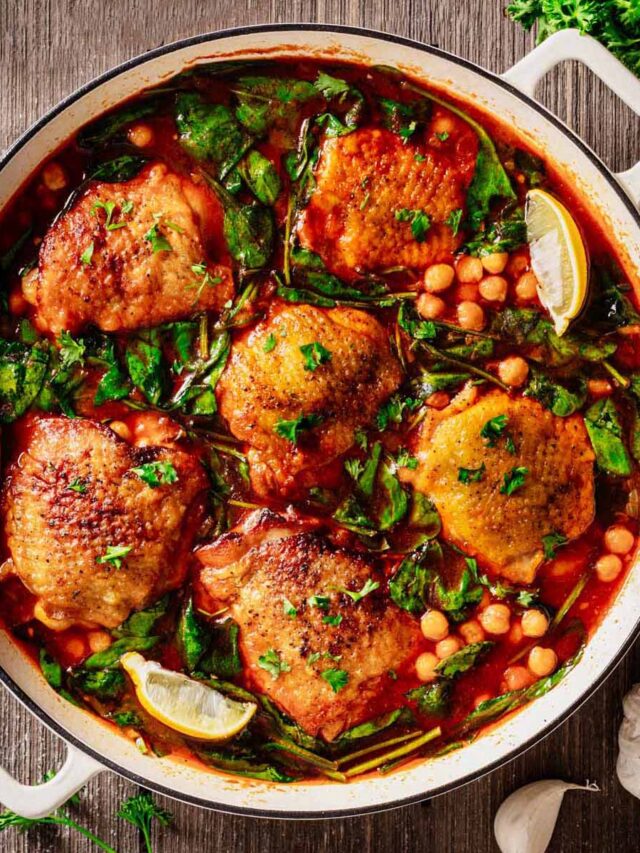 Mediterranean braised chicken in a white Dutch oven with golden-brown chicken thighs, garbanzo beans, spinach, and a rich tomato-based sauce, garnished with parsley and lemon wedges.