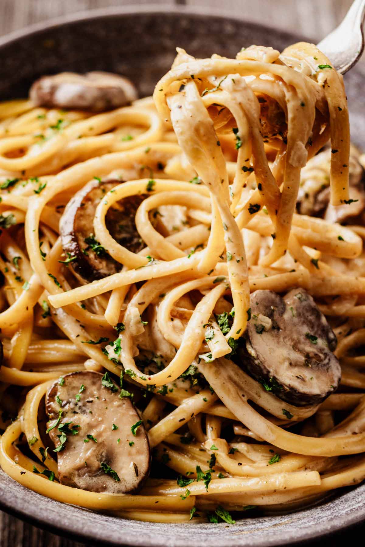 Close-up of a fork twirling creamy mushroom pasta with sliced mushrooms and parsley garnish in a gray bowl.