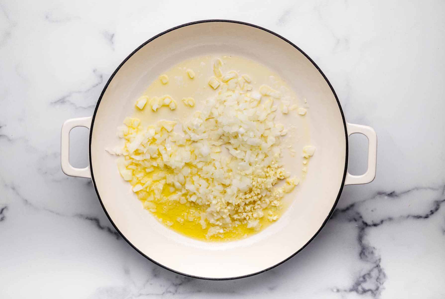 Chopped onions and minced garlic sautéing in melted butter and olive oil in a large white skillet.