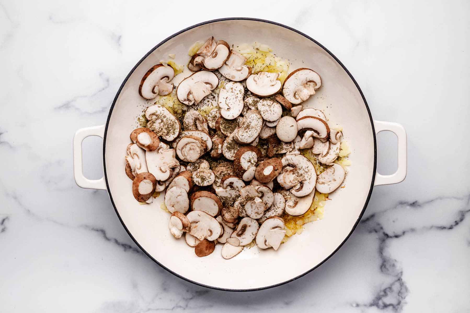 Sliced mushrooms seasoned with dried thyme, salt, and pepper added to a skillet with sautéed onions and garlic.