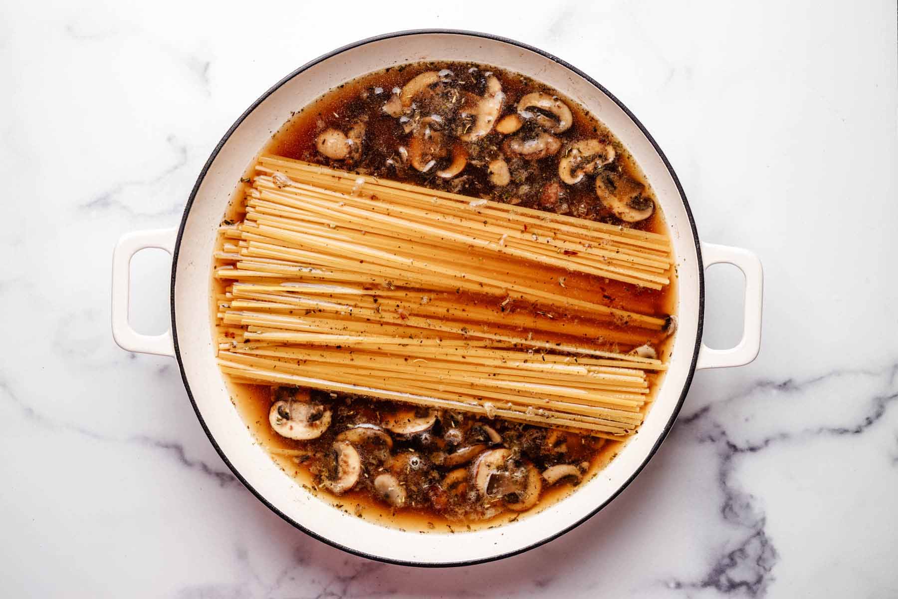 Dry linguine pasta placed in a pot with sliced mushrooms and seasoned broth, ready to cook for a one-pot mushroom pasta recipe.