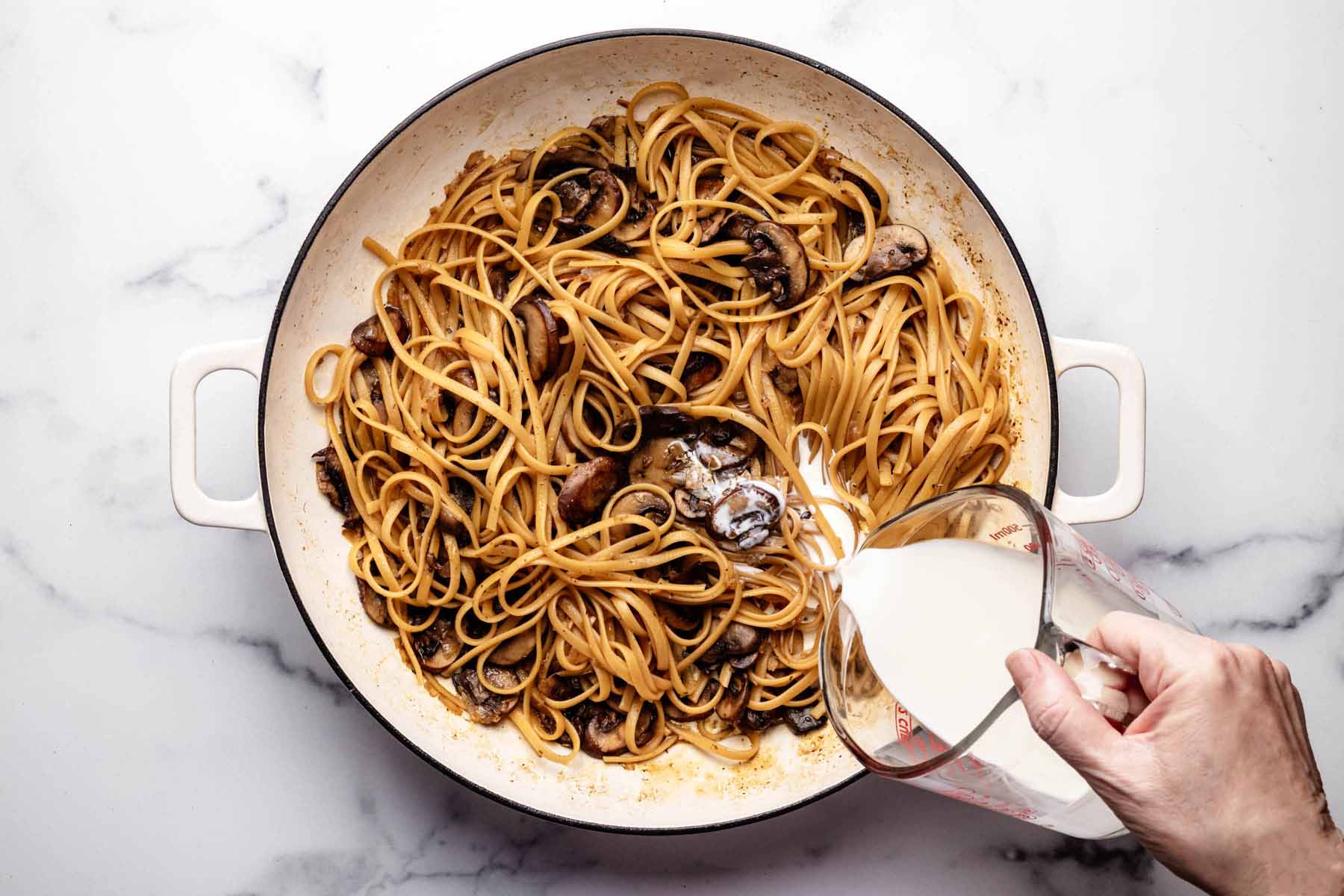 Cooked linguine and mushrooms in a large pot as heavy cream is poured in, creating the creamy sauce for a one-pot mushroom pasta recipe.