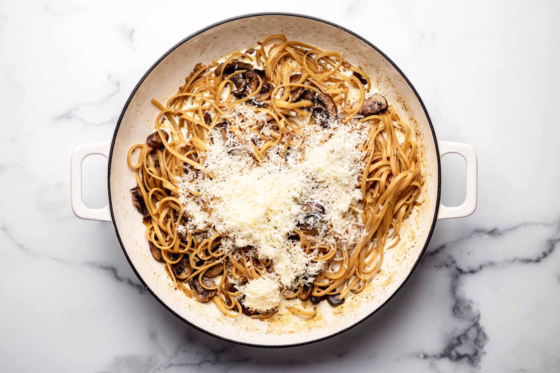 Linguine pasta and sautéed mushrooms in a large pot, topped with freshly grated Parmesan cheese, ready to be mixed into a creamy sauce.