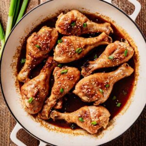Fully cooked braised chicken drumsticks coated in a rich soy-based sauce, garnished with sesame seeds and chopped green onions, served in a white enameled skillet on a wooden surface.