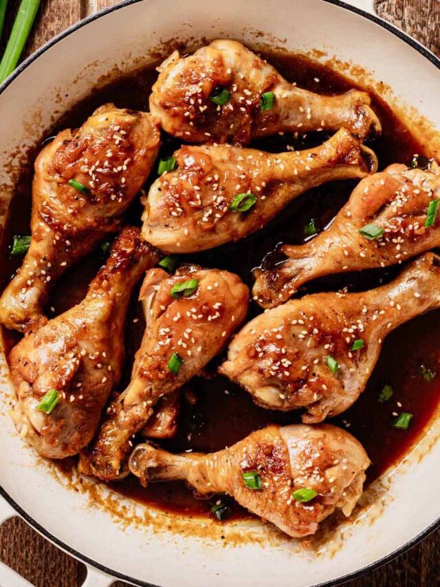 Fully cooked braised chicken drumsticks coated in a rich soy-based sauce, garnished with sesame seeds and chopped green onions, served in a white enameled skillet on a wooden surface.