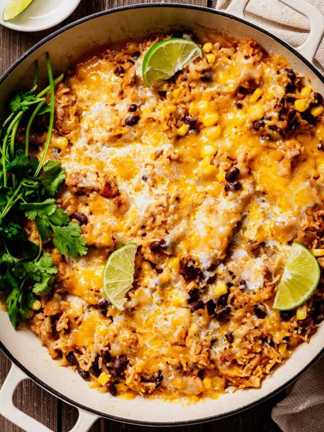 A skillet filled with Cheesy Mexican Chicken and Rice, topped with melted cheese, black beans, corn, fresh cilantro, and lime wedges, set on a rustic wooden surface.