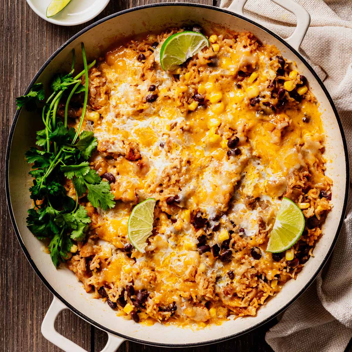 A skillet filled with Cheesy Mexican Chicken and Rice, topped with melted cheese, black beans, corn, fresh cilantro, and lime wedges, set on a rustic wooden surface.