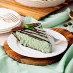 A slice of chocolate mint cheesecake with a creamy green filling, drizzled with dark chocolate ganache, and garnished with fresh mint. Served on a white plate with a fork, set on a wooden board with a mint-green napkin.