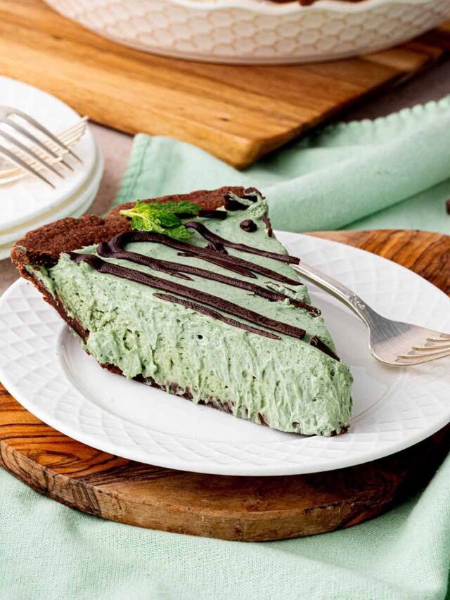 A slice of chocolate mint cheesecake with a creamy green filling, drizzled with dark chocolate ganache, and garnished with fresh mint. Served on a white plate with a fork, set on a wooden board with a mint-green napkin.