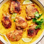 Golden-brown chicken thighs simmering in a creamy coconut milk sauce, garnished with lime wedges and fresh cilantro in a pan.