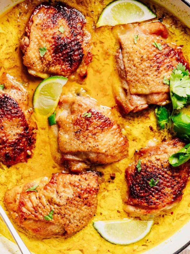 Golden-brown chicken thighs simmering in a creamy coconut milk sauce, garnished with lime wedges and fresh cilantro in a pan.