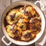 A white enameled cast iron skillet filled with French onion chicken thighs, topped with melted Swiss cheese, caramelized onions, and garnished with fresh parsley.