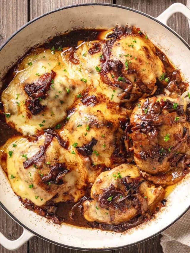 A white enameled cast iron skillet filled with French onion chicken thighs, topped with melted Swiss cheese, caramelized onions, and garnished with fresh parsley.