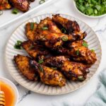 A plate of hot honey chicken wings with a caramelized glaze, garnished with sliced green onions. A bowl of extra green onions, a dish of hot sauce with a honey dipper, and a serving tray of more wings are arranged on a marble surface.
