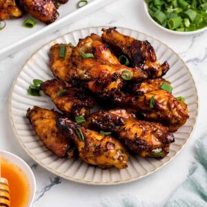 A plate of hot honey chicken wings with a caramelized glaze, garnished with sliced green onions. A bowl of extra green onions, a dish of hot sauce with a honey dipper, and a serving tray of more wings are arranged on a marble surface.