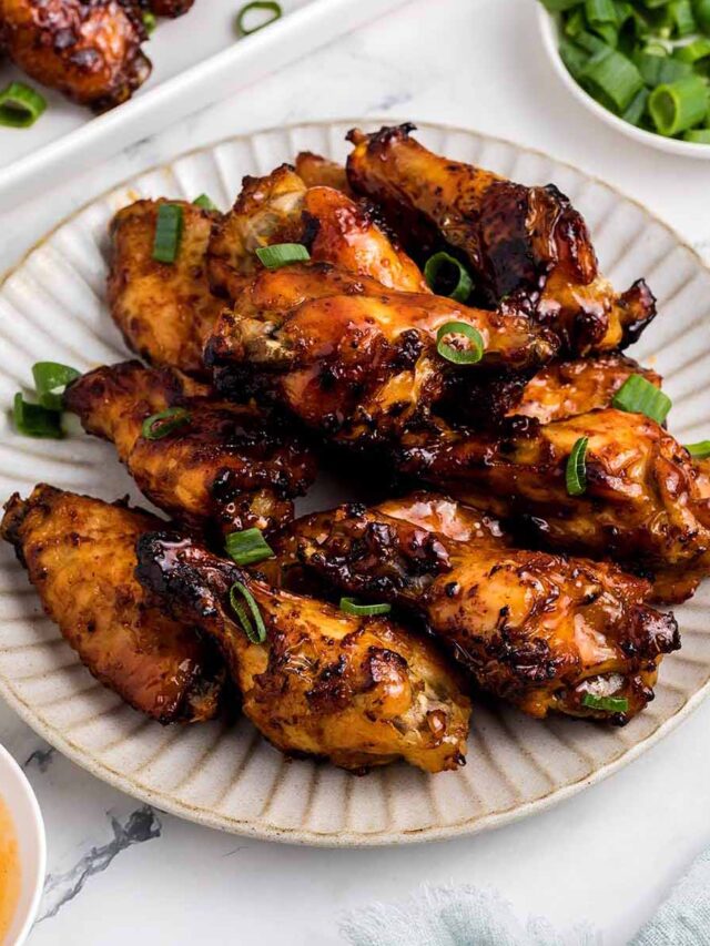 A plate of hot honey chicken wings with a caramelized glaze, garnished with sliced green onions. A bowl of extra green onions, a dish of hot sauce with a honey dipper, and a serving tray of more wings are arranged on a marble surface.
