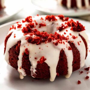 Close-up of a single mini red velvet bundt cake with cream cheese glaze and red velvet crumb topping on a white plate.