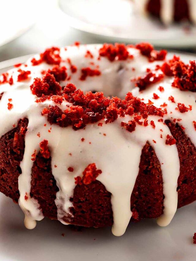 Close-up of a single mini red velvet bundt cake with cream cheese glaze and red velvet crumb topping on a white plate.