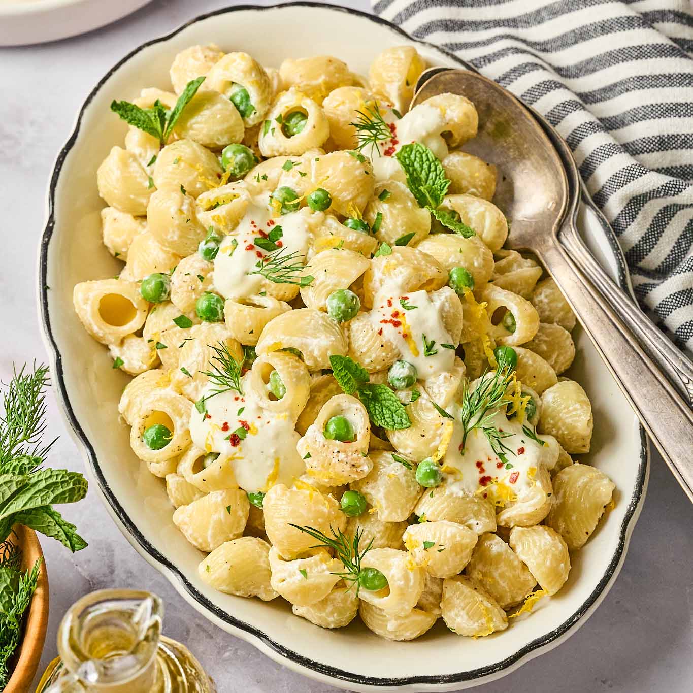 Creamy garlic lemon pasta with peas, garnished with mint, dill, lemon zest, and red pepper flakes, served in a white oval dish with a silver spoon and a striped napkin on the side.