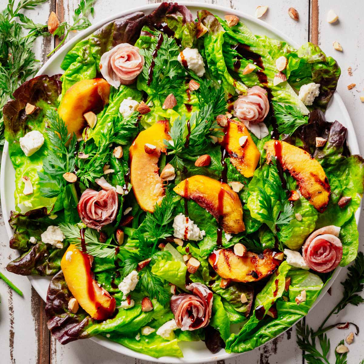 Grilled Peach & Prosciutto Salad with caramelized peaches, prosciutto rosettes, Boursin cheese, toasted almonds, and fresh mixed greens drizzled with balsamic glaze, served on a white plate.