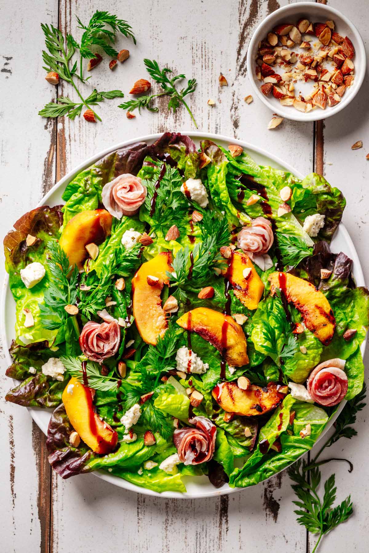 Grilled Peach & Prosciutto Salad with caramelized peaches, prosciutto rosettes, Boursin cheese, toasted almonds, and fresh mixed greens drizzled with balsamic glaze, served on a rustic white plate.