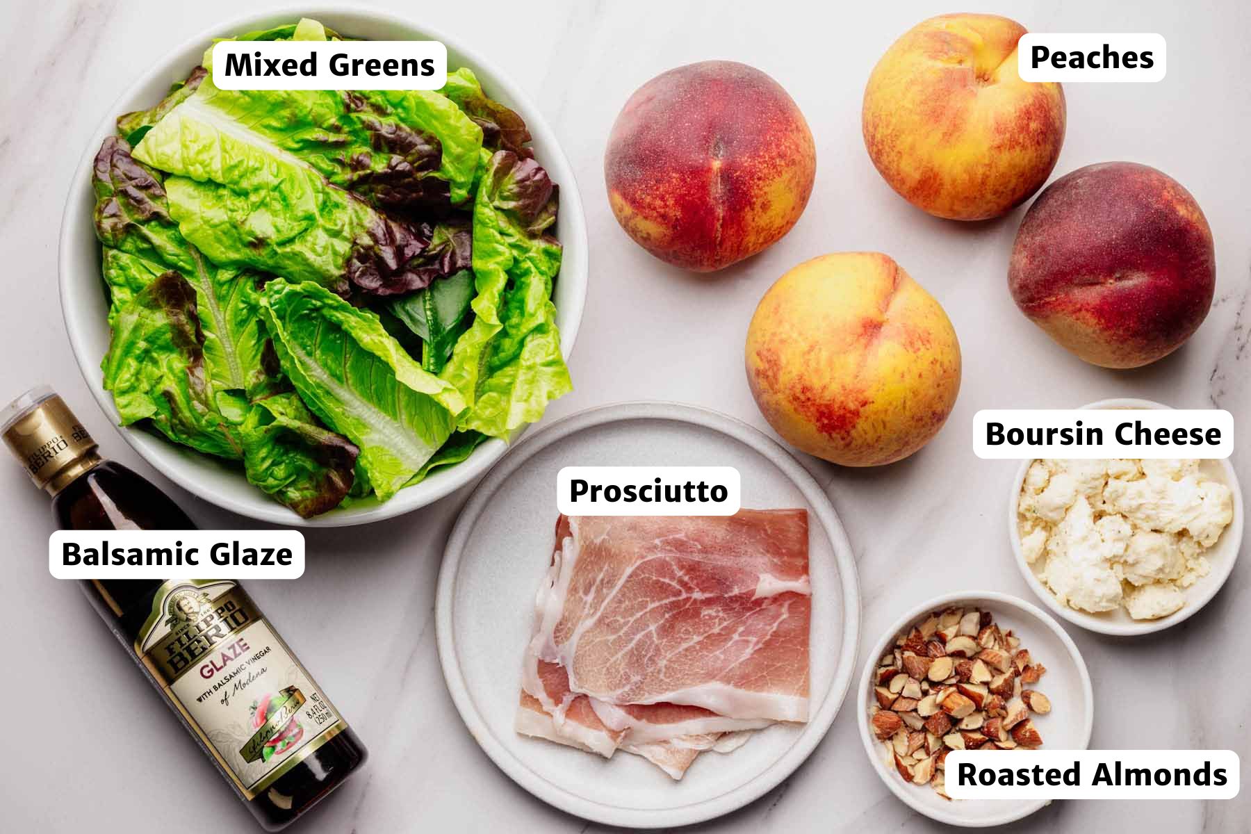 Labeled ingredients for grilled peach & prosciutto salad on a white marble surface: mixed greens, peaches, prosciutto, Boursin cheese, roasted almonds, and balsamic glaze.