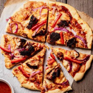 Sliced jerk chicken pizza with BBQ sauce, red onion, red bell pepper, and mango chipotle glaze on parchment paper, with a bowl of extra sauce on the side.