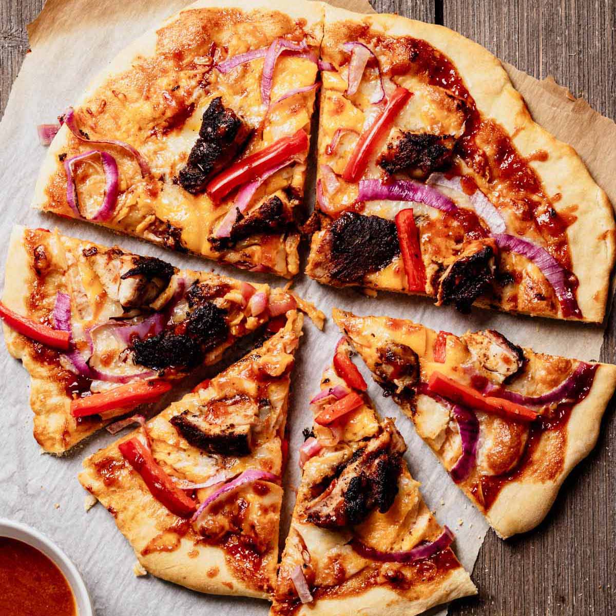 Sliced jerk chicken pizza with BBQ sauce, red onion, red bell pepper, and mango chipotle glaze on parchment paper, with a bowl of extra sauce on the side.