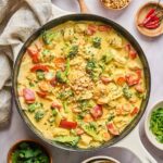 Yellow curry chicken with red bell peppers, broccoli, and carrots in a creamy curry sauce, garnished with chopped peanuts and fresh cilantro, served in a cream-colored cast iron skillet.
