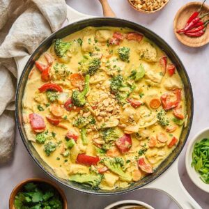 Yellow curry chicken with red bell peppers, broccoli, and carrots in a creamy curry sauce, garnished with chopped peanuts and fresh cilantro, served in a cream-colored cast iron skillet.
