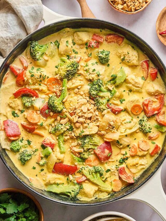 Yellow curry chicken with red bell peppers, broccoli, and carrots in a creamy curry sauce, garnished with chopped peanuts and fresh cilantro, served in a cream-colored cast iron skillet.