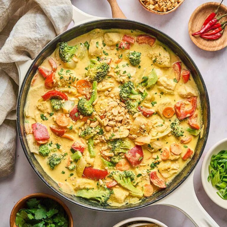 Yellow curry chicken with red bell peppers, broccoli, and carrots in a creamy curry sauce, garnished with chopped peanuts and fresh cilantro, served in a cream-colored cast iron skillet.