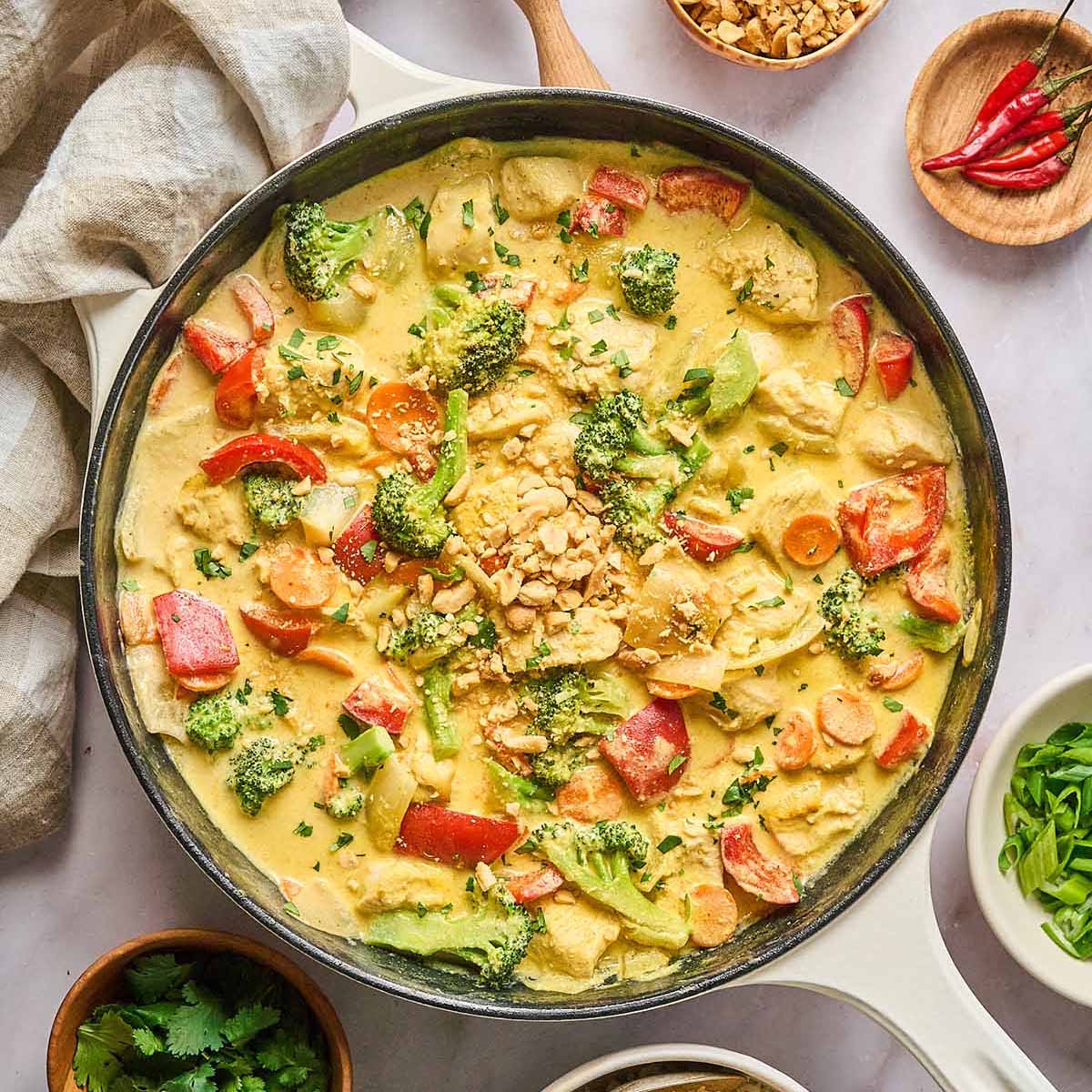 Yellow curry chicken with red bell peppers, broccoli, and carrots in a creamy curry sauce, garnished with chopped peanuts and fresh cilantro, served in a cream-colored cast iron skillet.