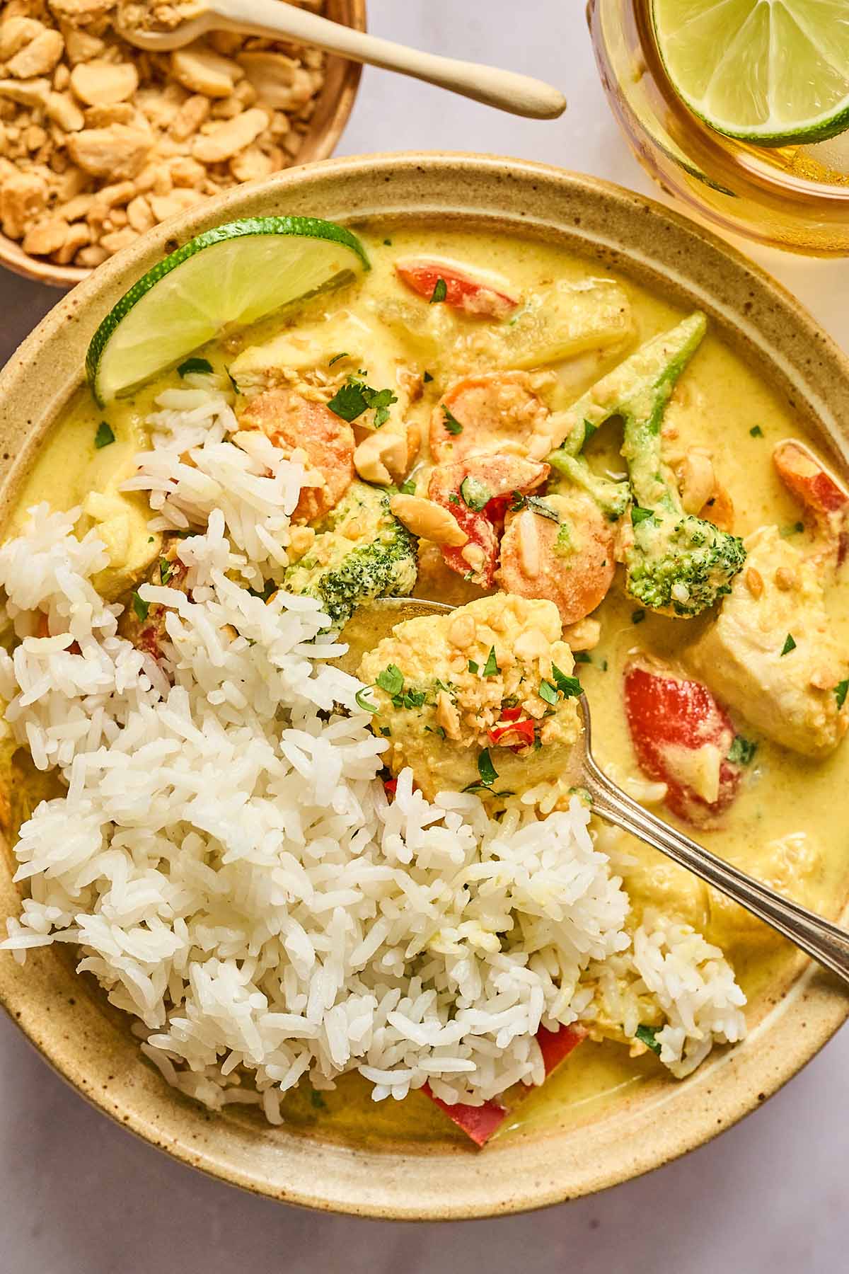 Bowl of yellow curry chicken with red bell peppers, broccoli, and carrots in a creamy curry sauce, served with white rice and garnished with chopped peanuts, cilantro, and a lime wedge.