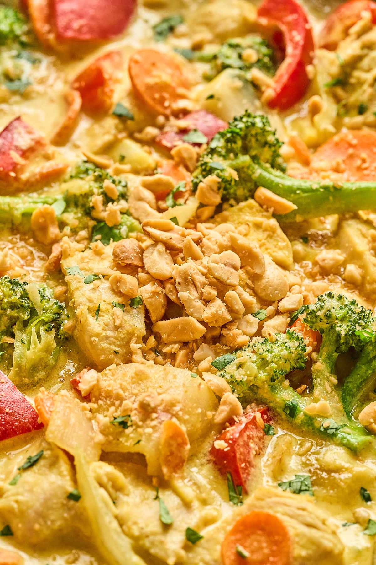 Close-up of yellow curry chicken with red bell peppers, broccoli, and carrots, garnished with chopped peanuts and fresh cilantro.