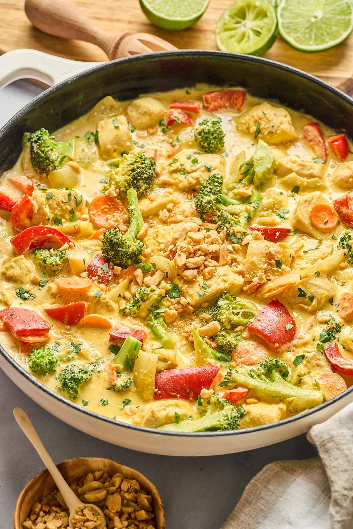 Yellow curry chicken with red bell peppers, broccoli, and carrots in a creamy curry sauce, garnished with chopped peanuts and fresh cilantro, with lime wedges and extra peanuts in the background.