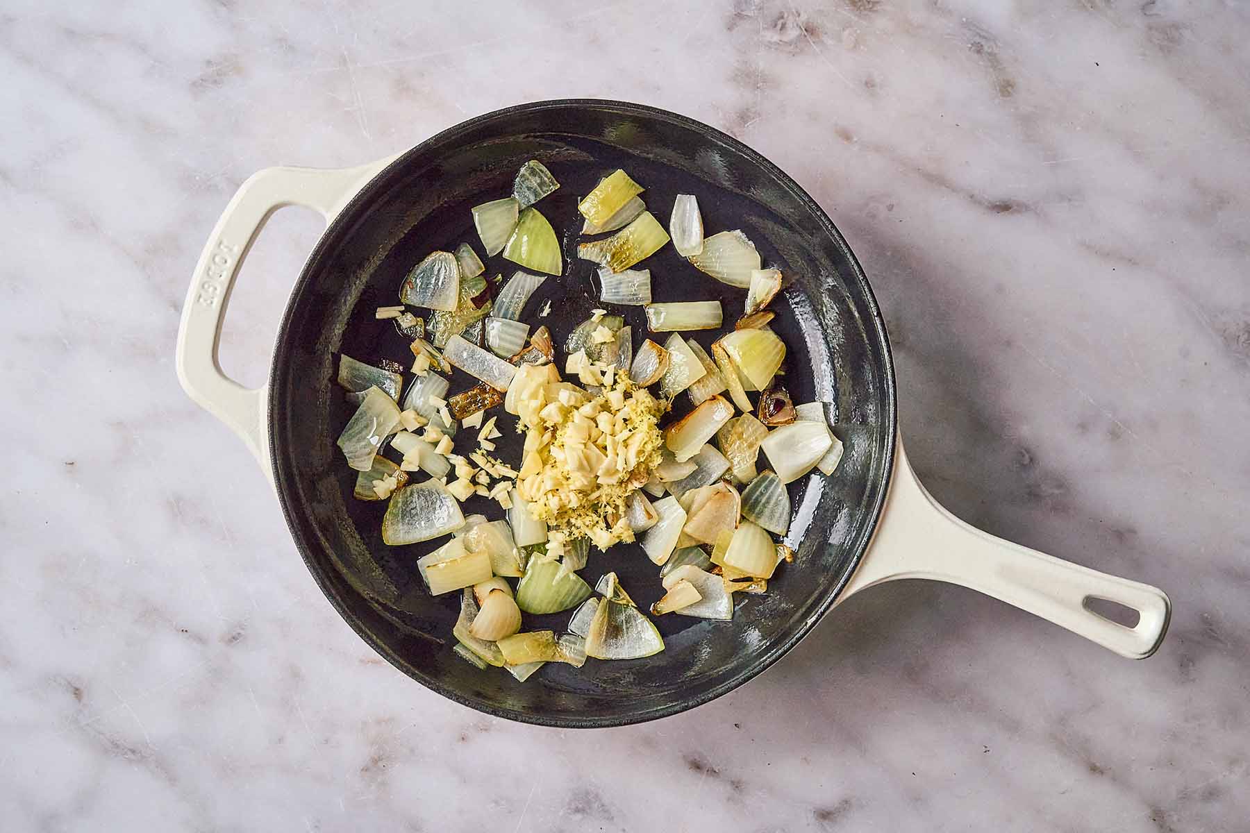 Chopped onions sautéing in a cream-colored cast iron skillet with minced garlic and grated ginger added to the center.