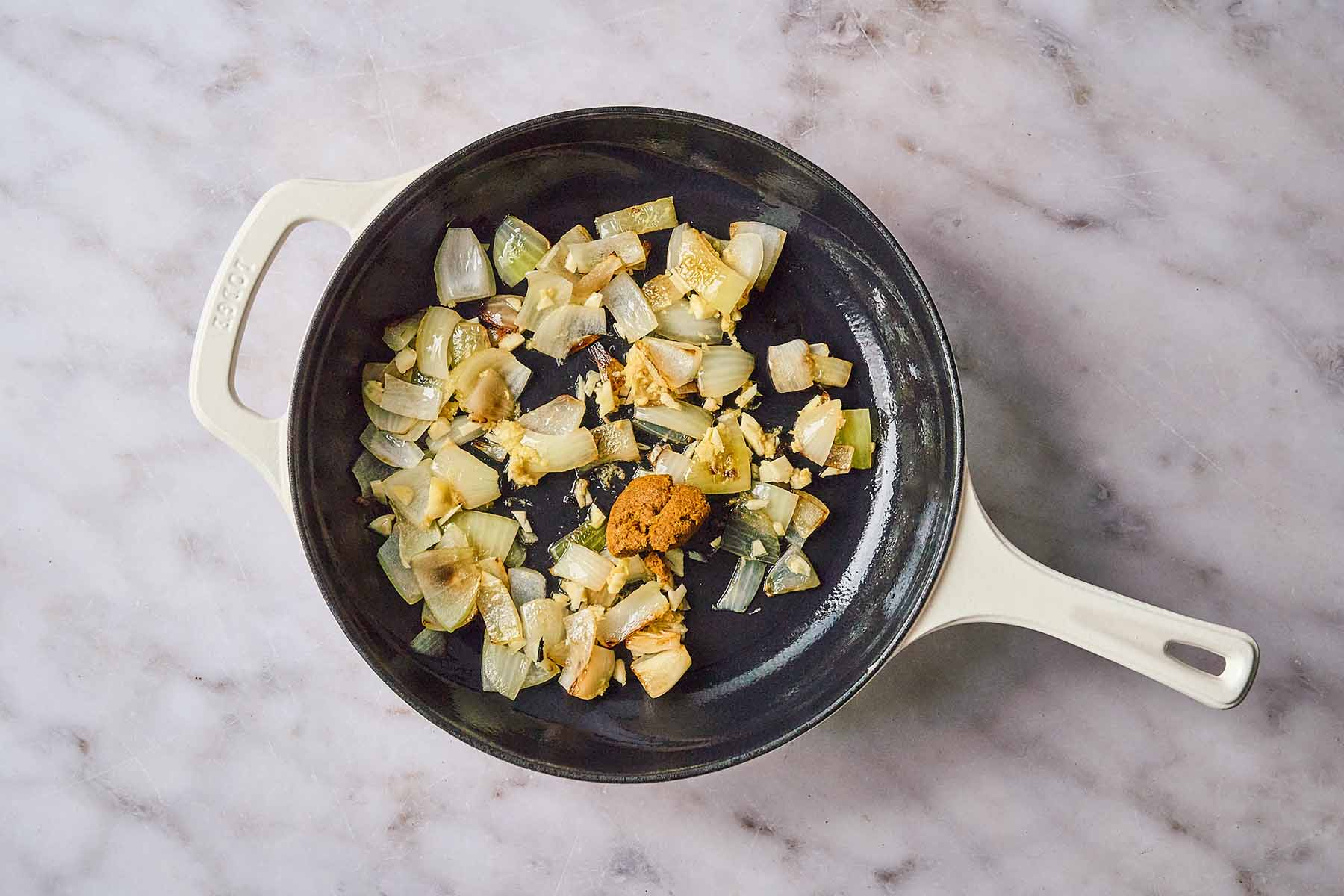 Chopped onions, garlic, and ginger sautéing in a cream-colored cast iron skillet with yellow curry paste added to the center.