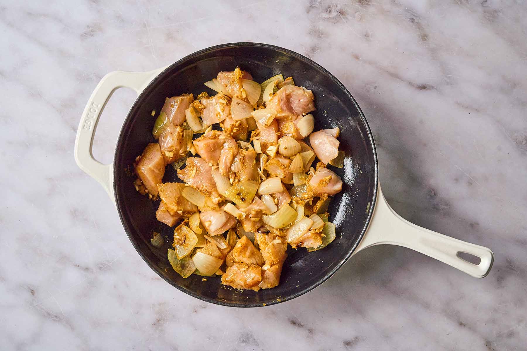 Raw chicken pieces added to sautéed onions, garlic, ginger, and curry paste in a cream-colored cast iron skillet.