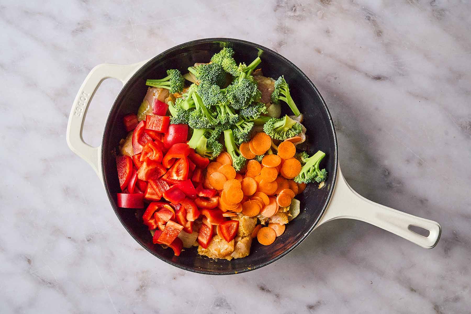 Chopped red bell peppers, broccoli florets, and sliced carrots added to chicken and sautéed aromatics in a cream-colored cast iron skillet.