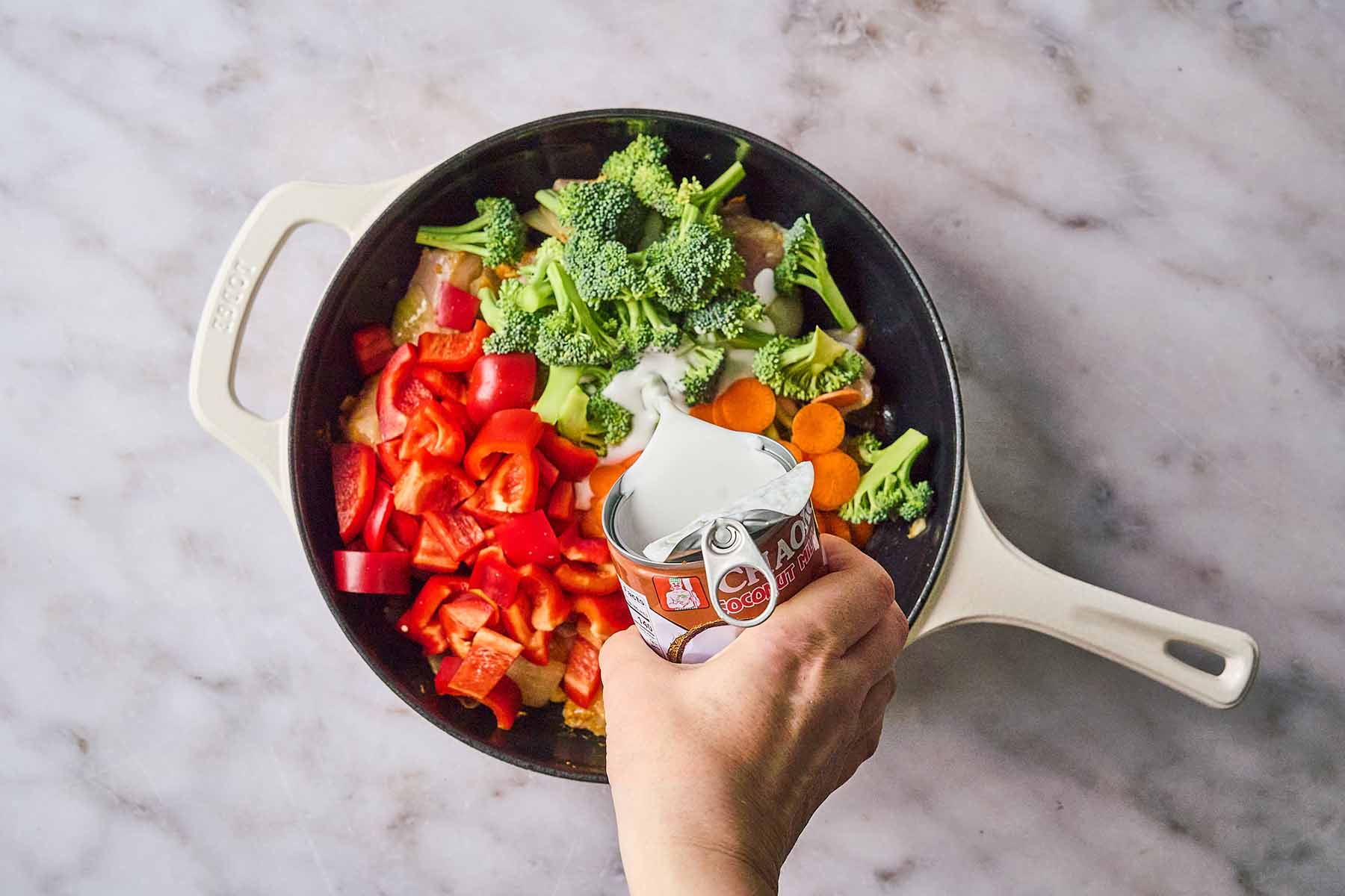 A hand pouring coconut milk from a can into a skillet filled with chicken, red bell peppers, broccoli florets, and sliced carrots.